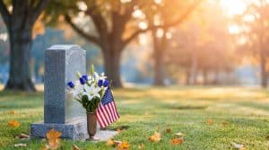 A gently decorated grave with fresh seasonal flowers in a flush vase and a small flag beside a headstone, in warm ivory and deep blue tones
